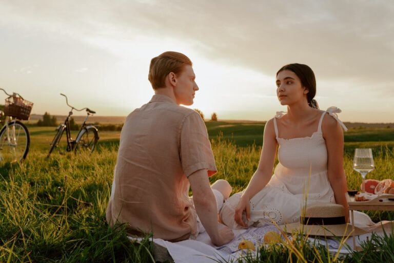 A couple sits on a picnic blanket in a sunny field, sharing a quiet moment. Two bicycles are nearby, and a sunset creates a warm, serene atmosphere.