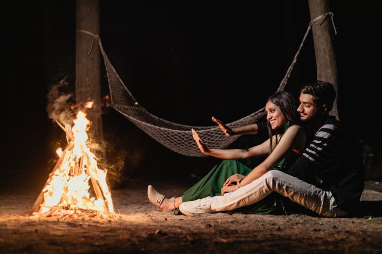 A couple sits by a campfire in the woods at night, warming hands, with a hammock in the background. The scene feels cozy and intimate.