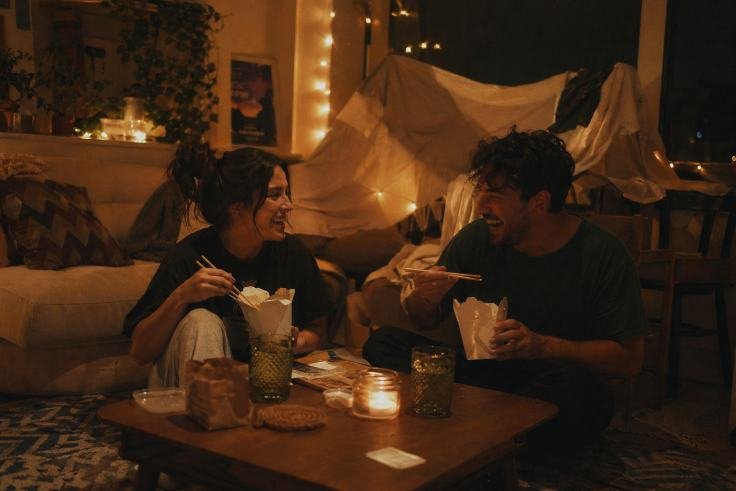 A couple sits on the floor, joyfully eating takeout with chopsticks in a cozy, warmly lit living room. Candles and string lights create a relaxed ambiance.