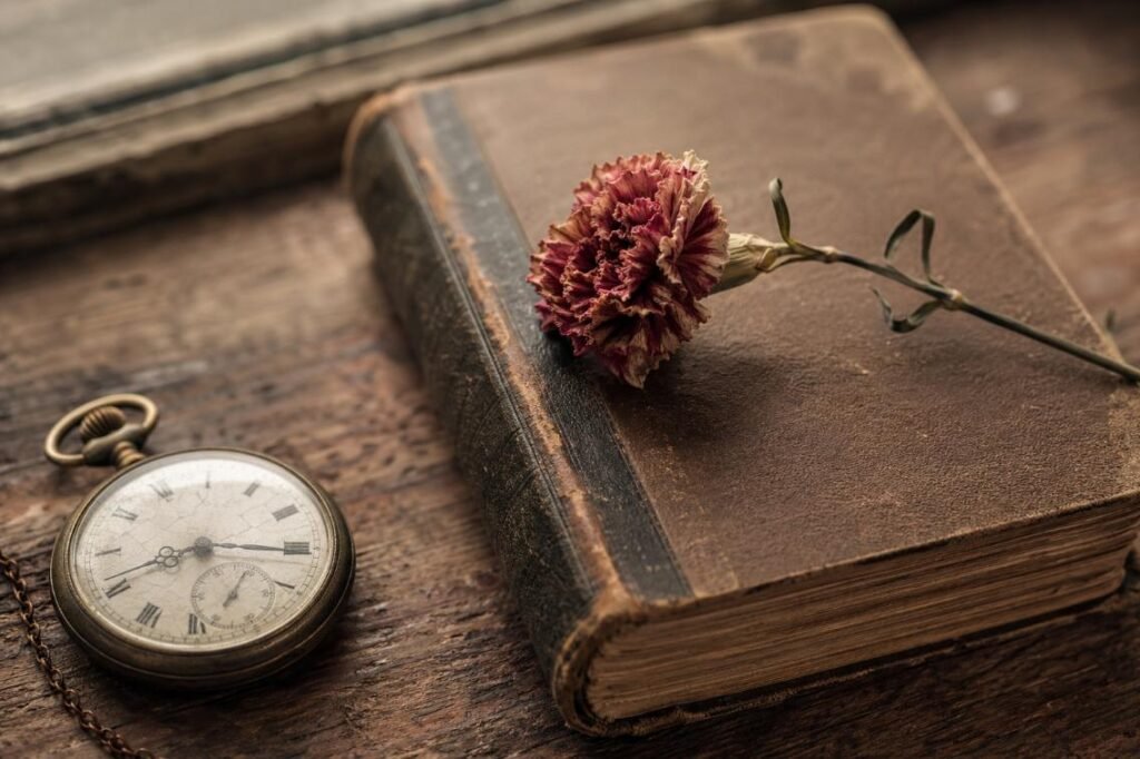 A vintage pocket watch rests beside a worn book with a dried red carnation on top, evoking a sense of nostalgia and timelessness on a wooden surface.