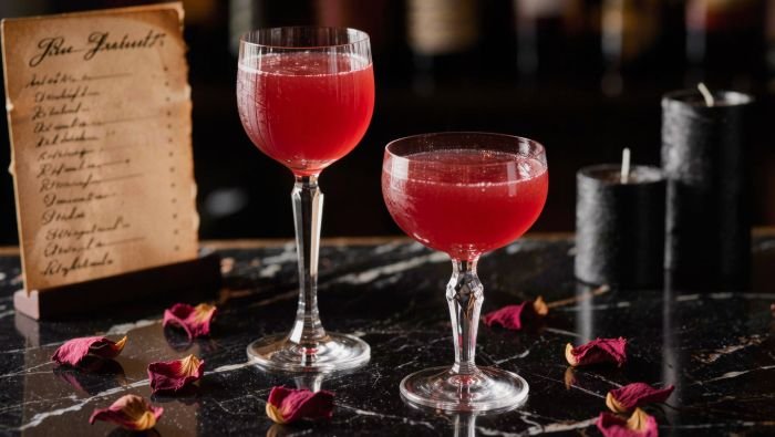 Two deep red cocktails in mismatched vintage glassware, dried rose petals scattered on a dark marble surface, black candles, an old handwritten cocktail menu card, moody overhead lighting, close-up editorial style