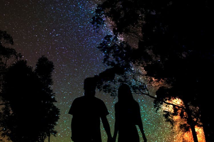 A silhouetted couple holds hands under a vibrant, starry night sky. The Milky Way glows with swirling colors, framed by dark tree outlines. Romantic and serene.