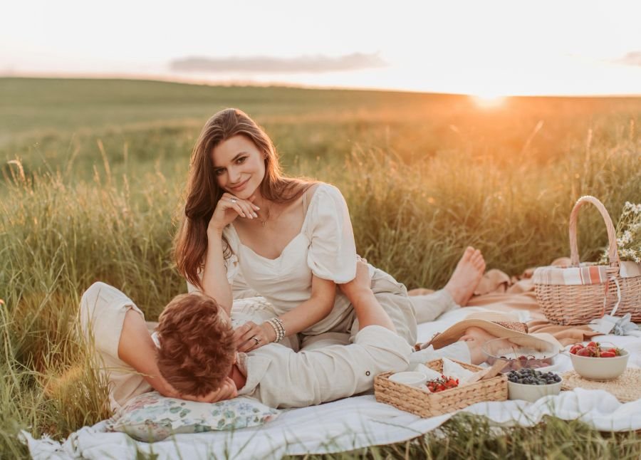A couple enjoys a serene picnic in a grassy field at sunset. The woman, in a white dress, smiles gently while the man rests his head on her lap. Baskets of fruit and a hat are by their side, creating a peaceful and romantic scene.