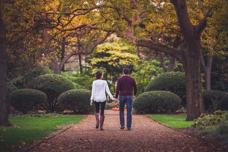 A couple walks hand in hand down a tree-lined path in an autumn garden, surrounded by lush green and orange foliage, creating a peaceful, romantic scene.
