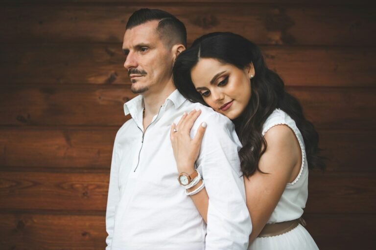 Couple in white clothing embracing against wooden wall.