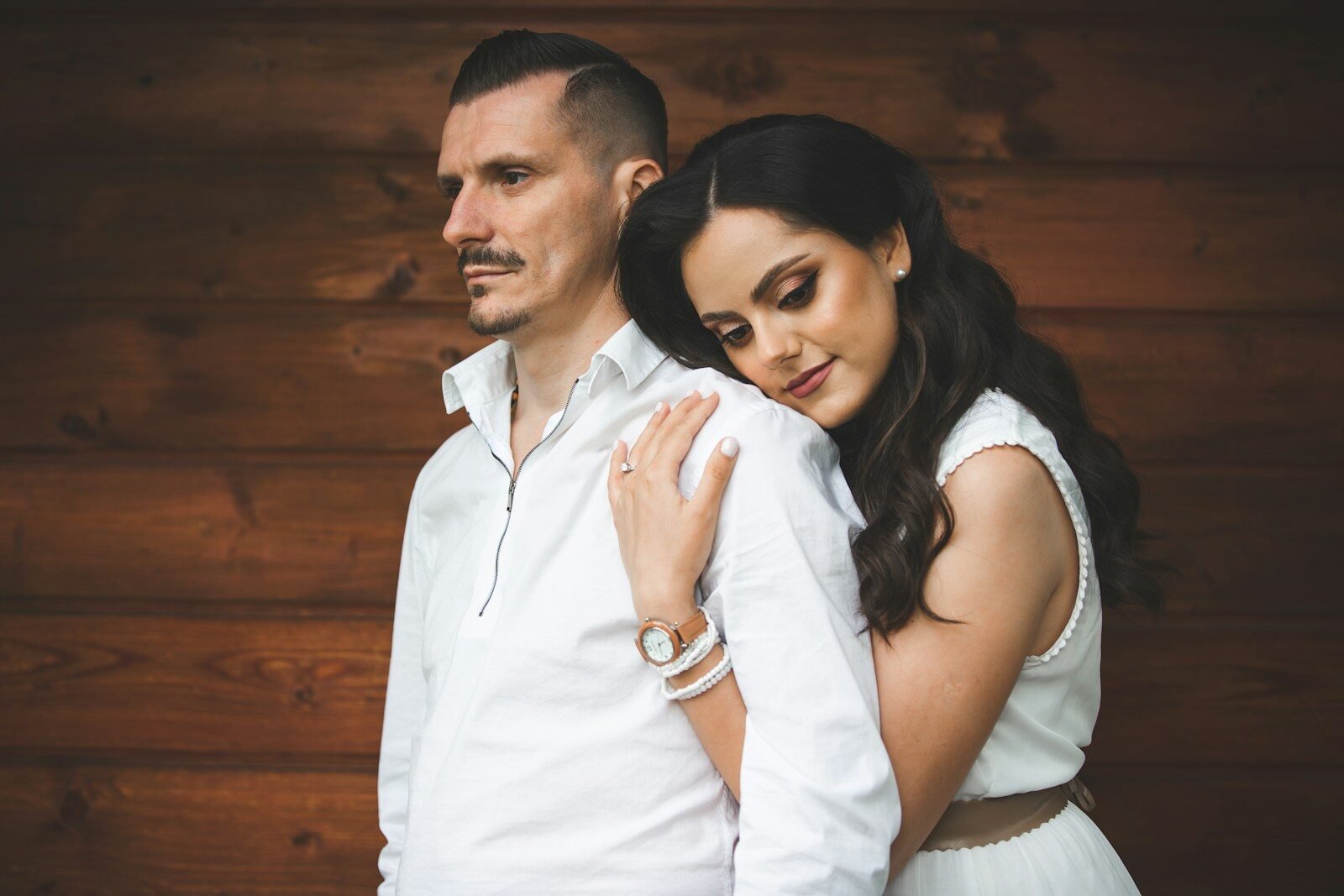 Couple in white clothing embracing against wooden wall.