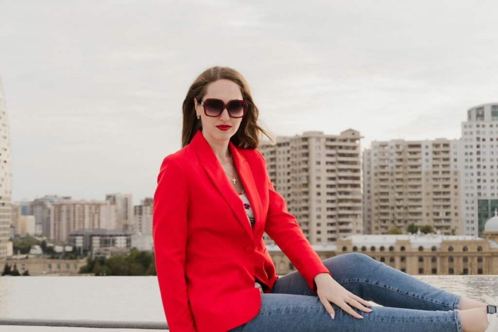 Woman in red blazer sitting on rooftop with city buildings.