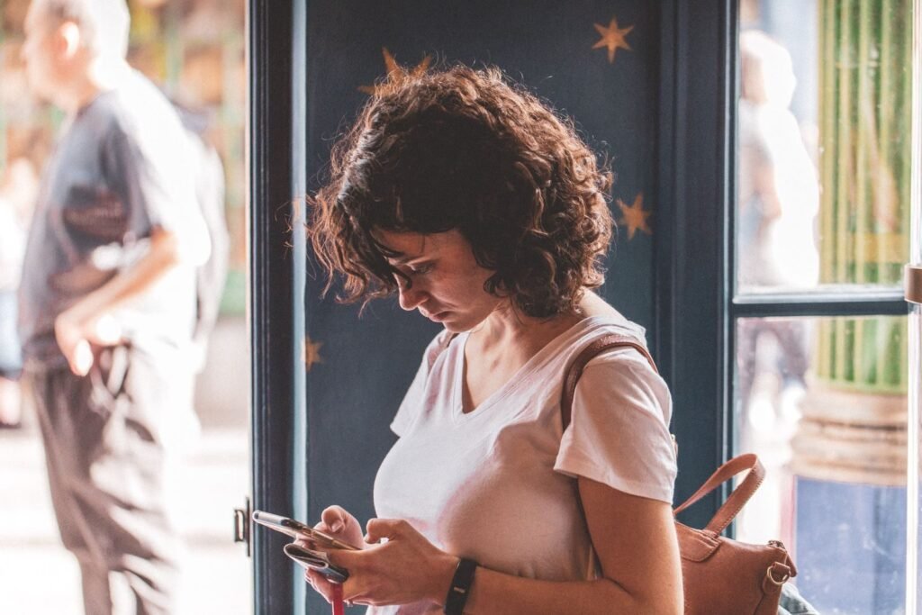 Woman with curly hair using smartphone by a sunlit window, wearing a white t-shirt.