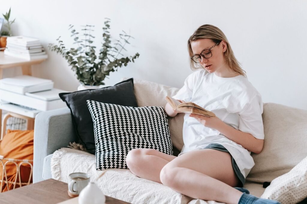 Serious young female in eyeglasses reading book on couch placed near table with cup of hot drink